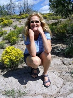 A color photograph of memoir editor and author Lisa Dale Norton while on a hike in the mountains outside Santa Fe, New Mexico. She is wearing blue shorts, a blue-and-white stripped short-sleeved top, and sandals. She is crouching over her heels with elbows on her knees smiling broadly at the camera. She sports John Lennon-like sunglasses. Her long, reddish-blonde hair flows over her shoulders. Around her bunches of yellow flowers bloom amidst pinion pines and juniper trees. Blue skies and puffy white clouds fill out the horizon. Norton is one of America's foremost independent editors of memoir. She is a gifted story analyst and a compassionate and wise teacher of the art and craft of writing. Norton is the author of America's favorite handbook for writing memoir entitled Shimmering Images: A Handy Little Guide to Writing Memoir.